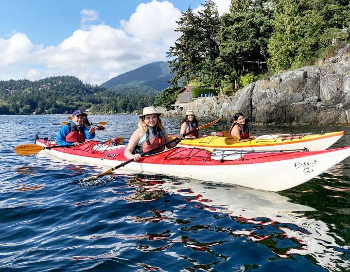 Four students kayaking in pairs on a crystal clear lake behind a natural landscape on a sunny day