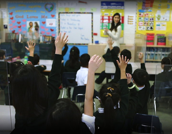 students with raised hands sitting in front of a teacher by a white board