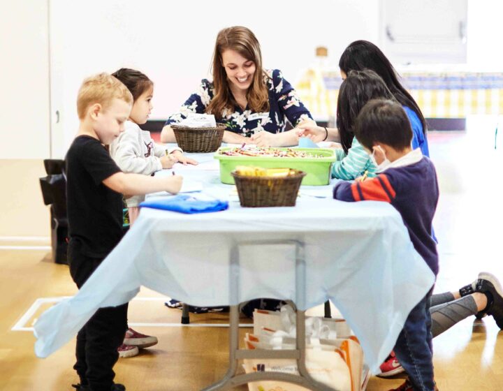 Teacher and five primary students colouring with crayons on a long table in the gym.