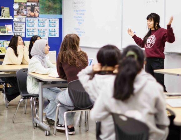 Teacher standing in front of a wide board holding a lesson for students sitting at their desks.