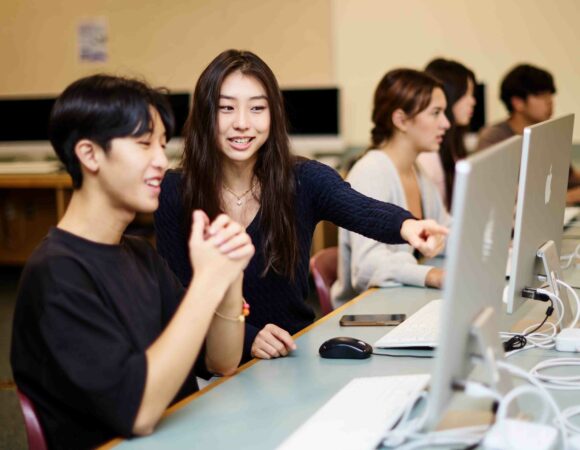 Female secondary student pointing at a computer screen sitting next to a male student who is smiling.