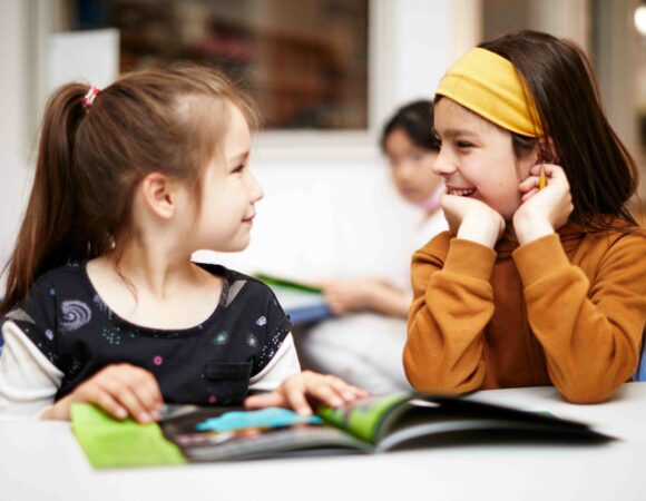 Two primary aged children reading a book and smiling at one another.