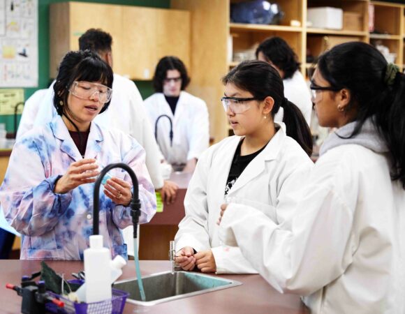 Students and teachers in lab coats conducting science experiment. One student is holding a test tube.