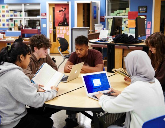 Group of secondary students sitting around a table. Some students are reading books while the others are looking at their computer screens.